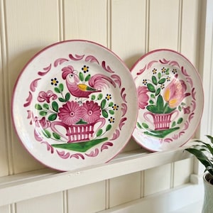 Könnte beinhalten: Zwei runde Keramikteller mit cremefarbener Basis und rosa Rändern. Der größere Teller zeigt einen handgemalten rosa Hahn über einem rosa Blumenkorb. Der kleinere Teller zeigt ein Arrangement aus rosa und gelben Tulpen in einem Korb.