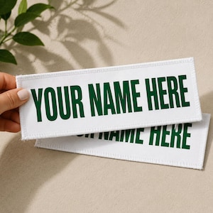 Könnte beinhalten: Zwei weiße rechteckige Aufnäher mit dem Text "YOUR NAME HERE" in grünen Blockbuchstaben. Die Aufnäher haben einen genähten Rand. Der Hintergrund ist beige.