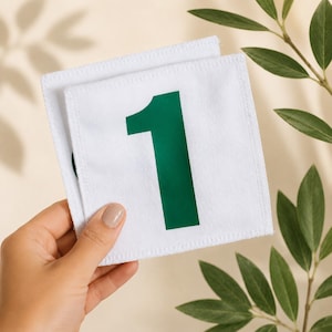 Pode incluir: Duas peças quadradas de tecido branco com o número 1 verde impresso. As peças são seguradas por uma mão, com folhas verdes ao fundo. As peças têm uma borda costurada.