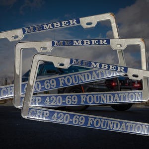 Op de afbeelding: Verchroomde kentekenplaathouders met de tekst "MEMBER" en "CHP 420-69 FOUNDATION" in blauwe letters. De frames zijn gestapeld voor een voertuig, met een bewolkte lucht op de achtergrond.