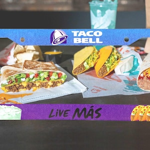 May include: A blue license plate frame featuring the Taco Bell logo and various menu items. The frame has the words "TACO BELL" and "LIVE MÁS" printed on it. The image includes a quesadilla, tacos, and a burrito.