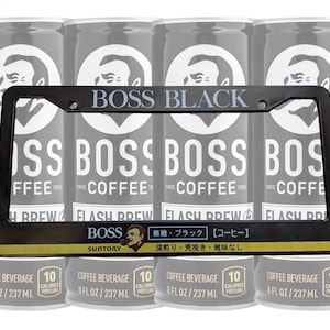 Könnte beinhalten: Ein schwarzer Kennzeichenrahmen mit dem Text "BOSS BLACK" und "BOSS COFFEE" in weißer Schrift. Hinter dem Rahmen befinden sich vier Dosen BOSS-Kaffee, jede mit einem Logo und Text. Jede Dose hat 237 ml.