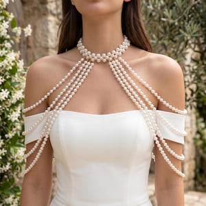 May include: A pearl body chain necklace with multiple strands cascading over the shoulders. The necklace is composed of small, round, white pearls and is worn over a white off-the-shoulder dress. The background features a stone wall and greenery.