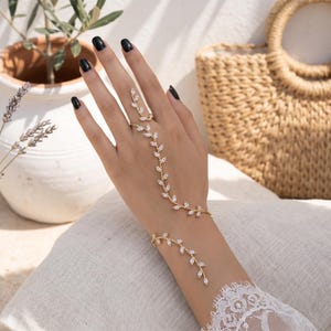 Puede incluir: Una cadena de mano dorada con un delicado diseño de enredadera y piedras transparentes en talla marquesa. La joya conecta un anillo en el dedo medio con una pulsera en la muñeca, creando un patrón continuo en forma de hojas.