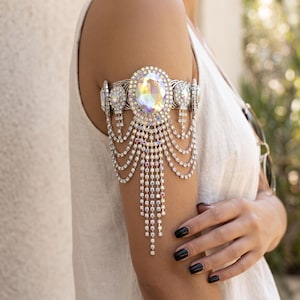 May include: An upper arm bracelet featuring a large central oval iridescent crystal surrounded by smaller clear rhinestones. The piece includes multiple draped silver-toned chains adorned with tiny sparkling crystals that hang down the arm.