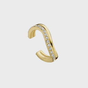 May include: A gold ear cuff with a wavy, open-ended design featuring a single row of small, clear, round-cut stones embedded along the center of the band. The metal has a polished, reflective finish, and the piece maintains a curved, fluid shape.