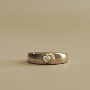 May include: A polished gold band ring featuring a single, small, heart-shaped setting at the center. The heart-shaped setting contains a tiny, clear, round-cut gemstone that is flush-mounted within the smooth, rounded surface of the metal band.