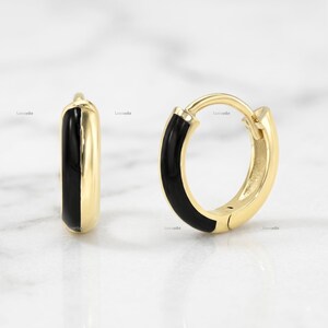 Puede incluir: Un par de pendientes de aro pequeños hechos de metal pulido en tono dorado, cada uno con una incrustación de esmalte negro liso y brillante en el borde exterior. Los pendientes tienen un mecanismo de cierre con bisagra y diseño circular.