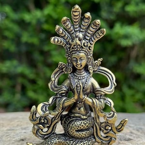 May include: A detailed brass statue of a deity with a multi-headed cobra headdress. The figure is in a seated position with hands clasped in prayer. The statue has intricate details and a weathered appearance, suggesting an antique or vintage piece.
