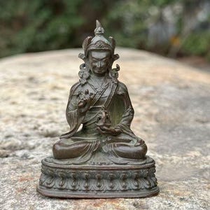 May include: A bronze-colored statue of a seated figure in a meditative pose. The figure wears a detailed headdress and robes, with hands in a symbolic gesture. The statue sits on a decorative base, showcasing intricate patterns.
