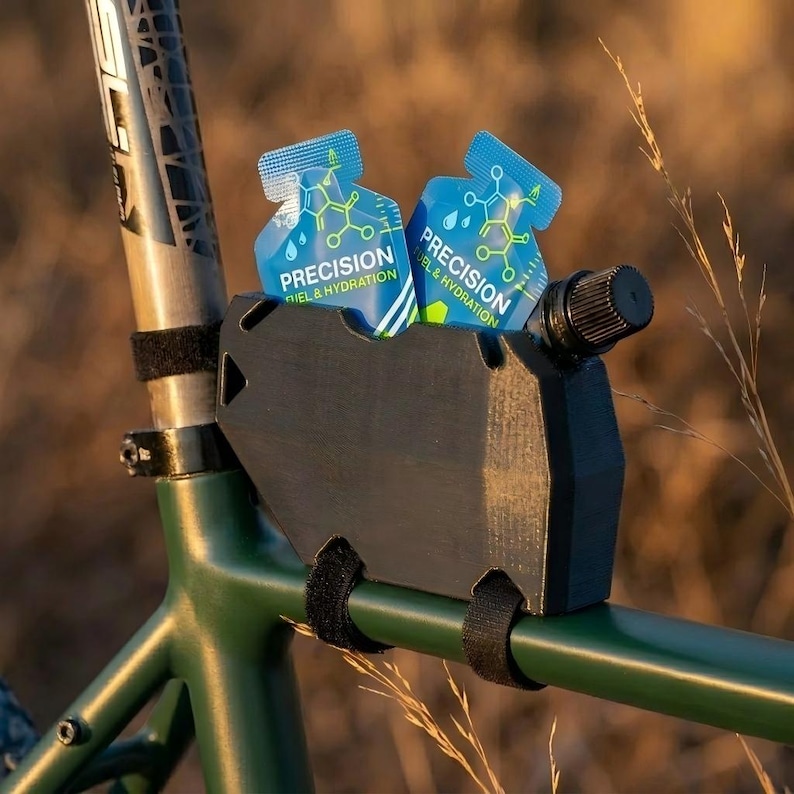 May include: A black bicycle accessory holds two blue and white packets labeled "Precision Fuel & Hydration." The accessory is mounted on a green bicycle frame with black velcro straps. The background is blurred with dry grass.