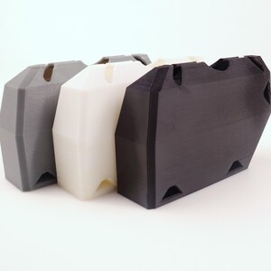 May include: Three 3D-printed storage containers in a row. The containers are in gray, white, and black. Each has a trapezoidal shape with a slot on top and triangular cutouts at the bottom corners. The containers are made of a matte plastic material.