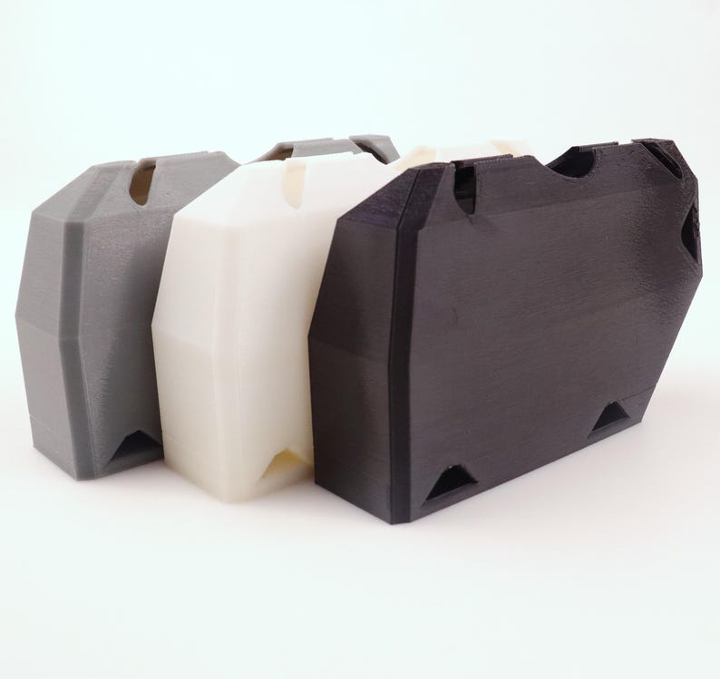 May include: Three 3D-printed storage containers in a row. The containers are in gray, white, and black. Each has a trapezoidal shape with a slot on top and triangular cutouts at the bottom corners. The containers are made of a matte plastic material.