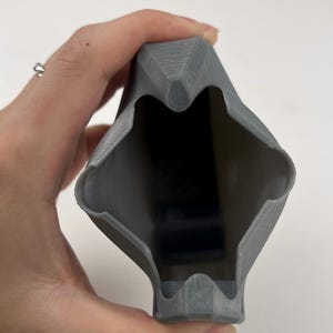 May include: A gray, 3D-printed object with a geometric design. The object has a hollow interior and is held in a hand. The exterior has a textured surface. The object's shape is angular and symmetrical.