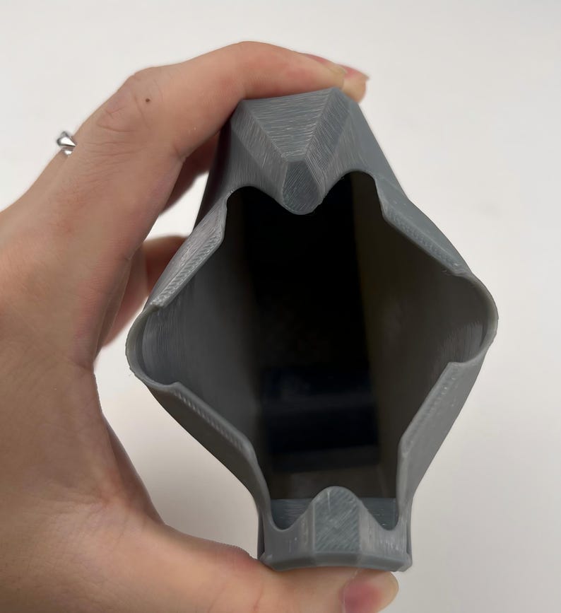 May include: A gray, 3D-printed object with a geometric design. The object has a hollow interior and is held in a hand. The exterior has a textured surface. The object's shape is angular and symmetrical.