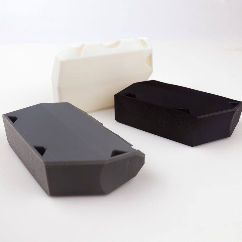 May include: Three rectangular, geometric objects in white, black, and gray. The objects have angled sides and small triangular cutouts. The white object has three curved indentations on top. The objects are arranged on a white surface.