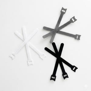 May include: An overhead shot displays several reusable cable ties in white, gray, and black. Each tie has a rectangular body with a slot and a tab at one end. The ties are arranged against a white background.