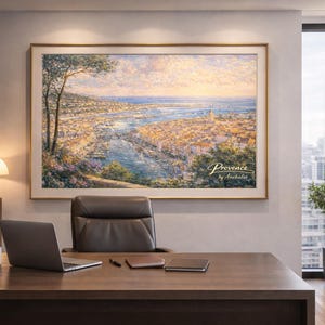May include: A framed landscape painting depicting a coastal city scene with a river and buildings. The artwork, labeled "Provence by Anchellee," hangs above a desk with a laptop, mug, and office supplies. A plant and a window with a city view are also visible.