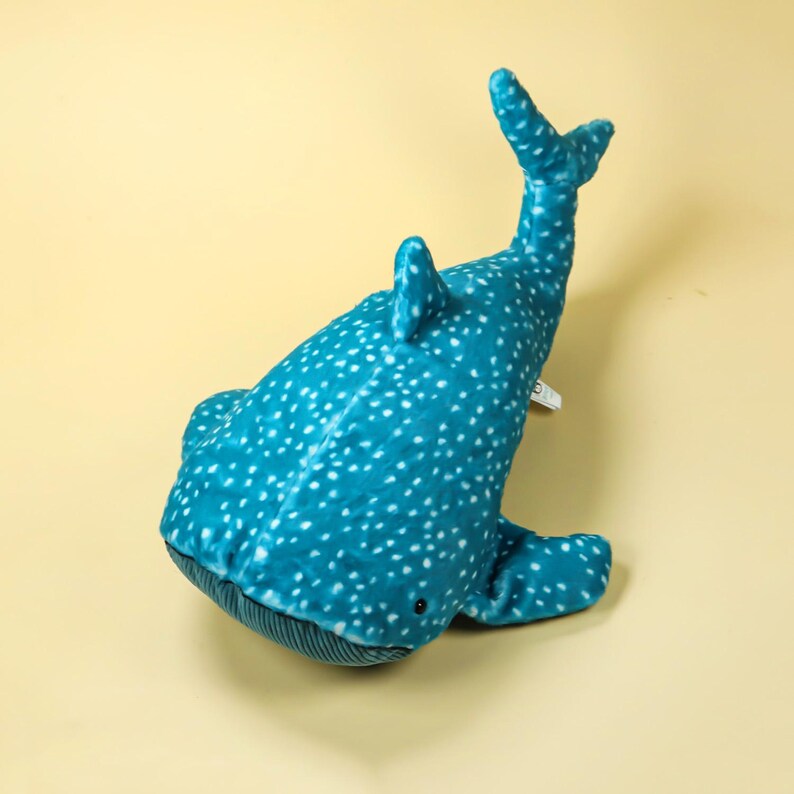Puede incluir: Un juguete de peluche de tibur&oacute;n ballena en tonos turquesa y azul, cubierto de puntos blancos. El juguete tiene un cuerpo redondeado, aletas y una cola, y est&aacute; dise&ntilde;ado para parecerse a la criatura marina. El juguete est&aacute; hecho de material suave.