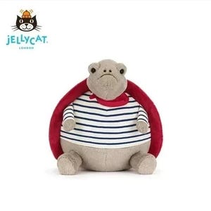Peut inclure: Une peluche en forme de tortue avec une tête et un corps gris, vêtue d'une chemise rayée bleue et blanche, d'une écharpe rouge et d'une carapace rouge. Le logo Jellycat est visible en haut à gauche.