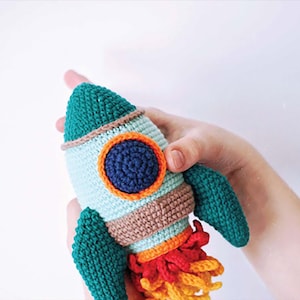 May include: A handmade crochet rocket ship toy in shades of teal, brown, orange, and red. The rocket has a teal cone-shaped top, a round blue window, and teal wings. The rocket's exhaust is made of red and yellow yarn.