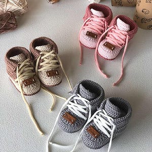 May include: Handmade crochet baby sneakers in three color combinations: pink and white, gray and white, and brown and beige. Each pair features laces and a small leather-like tag. The shoes are designed to resemble adult sneakers.