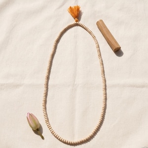 May include: A beige wooden bead necklace with an orange tassel, a wooden stick, and a pink flower bud on a beige fabric background. The necklace is made of small, round beads. The tassel is a bright orange color.