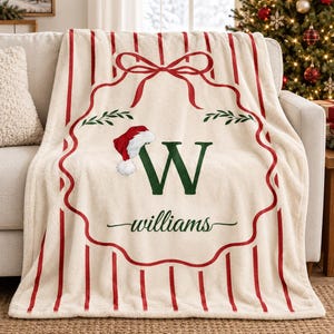 May include: A cream-colored fleece throw blanket featuring a red vertical striped pattern. The center displays a red outlined ornament shape with a bow, a green letter W, a Santa hat, and the text williams in green script lettering.
