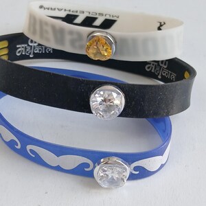 Puede incluir: Una pila de cuatro pulseras de goma. La pulsera superior es blanca con texto negro y una gema amarilla. La segunda pulsera es negra con texto hindi y una gema transparente. La tercera pulsera es azul con un diseño de bigote y una gema transparente.