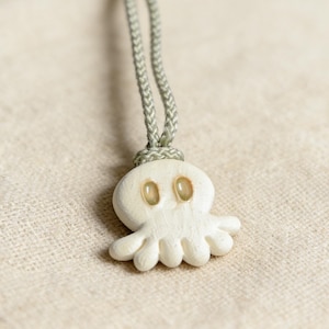 Puede incluir: Un colgante de pulpo color crema con un cordón verde claro. El pulpo tiene una cabeza redondeada, ocho tentáculos y dos ojos ovalados. El cordón está trenzado y anudado en la parte superior.