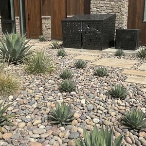 May include: A black metal HVAC privacy screen with a laser-cut geometric pattern, positioned on a stone-covered landscape featuring multiple agave plants, smooth river rocks, and light-colored rectangular stone pavers in front of a building.