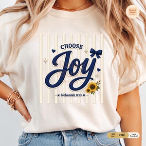May include: A cream-coloured t-shirt featuring a graphic design with vertical light stripes. The text reads CHOOSE Joy and Nehemiah 8:10 in dark blue script. The design includes a dark blue bow, small hearts, star accents, and two sunflowers.