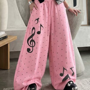 May include: Pink wide-leg sweatpants with a black polka dot pattern and musical note graphics. The pants have a drawstring waist and are worn with black platform shoes. The musical notes include a treble clef and eighth notes.