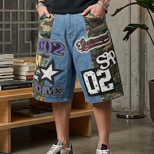 Puede incluir: Pantalones cortos de mezclilla con patrón de camuflaje, múltiples parches y texto gráfico que incluye 902, SP y 02. Los pantalones tienen un corte holgado, un gráfico de estrella blanca y la palabra ARCHIVES impresa en negrita en la pierna.