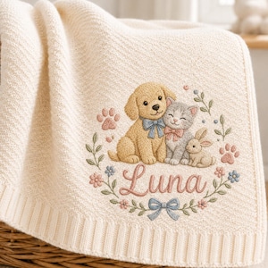 Pode incluir: Uma manta de malha cor de creme com um bordado que mostra um cachorro dourado, um gatinho cinzento e um coelho pequeno. Os animais estão rodeados por uma coroa de flores, pegadas e laços azuis. O nome Luna está bordado em cor-de-rosa.