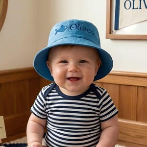 May include: A blue bucket hat featuring an embroidered fish icon and the name Oliver in dark blue script lettering. The hat has a wide, downward-sloping brim and is constructed from a solid blue fabric, providing a simple and functional design.