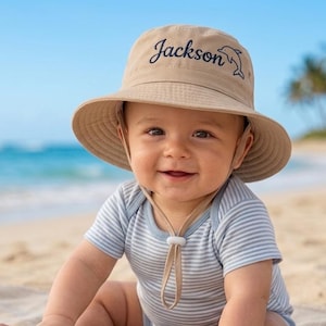 May include: A baby wearing a tan sun hat with the name "Jackson" embroidered on it, along with a dolphin graphic. The baby is wearing a blue and white striped onesie and is sitting on a beach with a toy turtle.