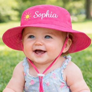 Peut inclure: Un chapeau de soleil rose vif avec le nom "Sophia" et un motif de soleil. Le chapeau a un large bord et une mentonnière. Le bébé porte un barboteuse bleue à fleurs et joue avec des jouets.