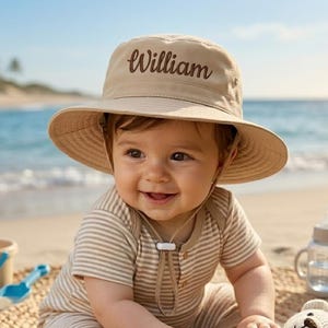 May include: A smiling baby wearing a tan sun hat with the name "William" embroidered on it. The baby is also wearing a striped onesie and sitting on the sand with a stuffed bear. A beach scene with seashells and a blue ocean in the background.