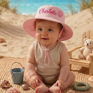 May include: A baby wearing a pink sun hat with the name "Charlotte" embroidered on it, sitting on a woven mat at the beach. The baby is wearing a beige onesie. A small bucket, seashells, and a teddy bear are nearby.