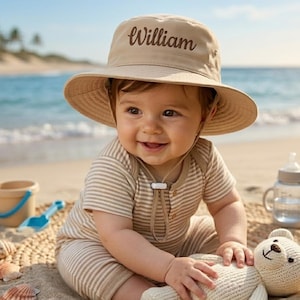 May include: A smiling baby wearing a tan sun hat with the name "William" embroidered on it. The baby is also wearing a striped onesie and sitting on the sand with a stuffed bear. A beach scene with seashells and a blue ocean in the background.