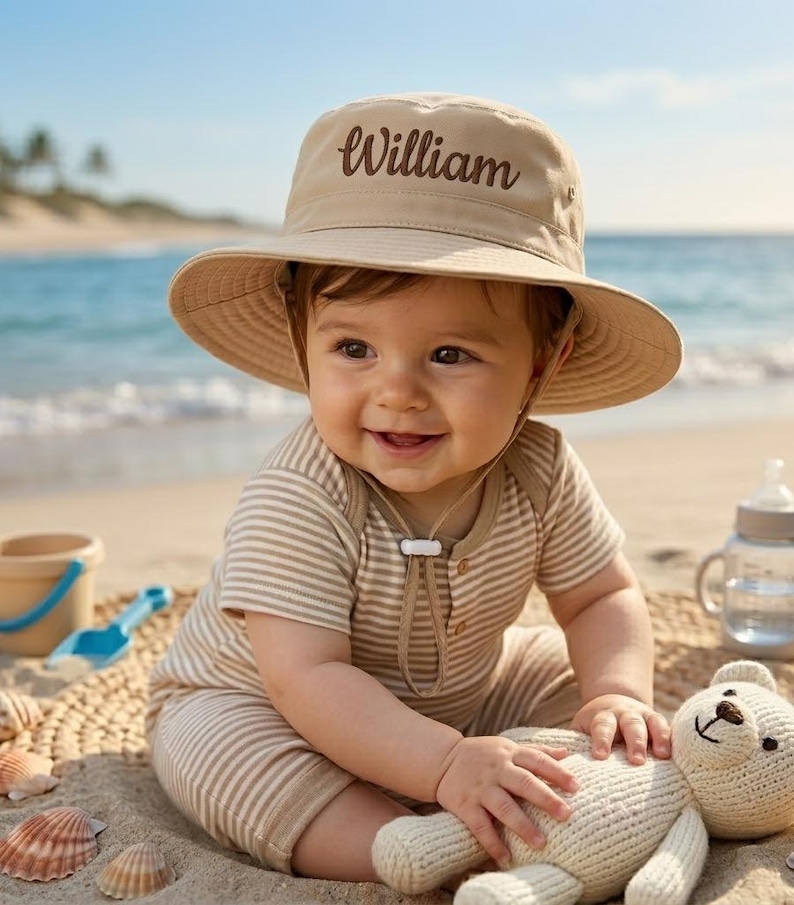 May include: A smiling baby wearing a tan sun hat with the name "William" embroidered on it. The baby is also wearing a striped onesie and sitting on the sand with a stuffed bear. A beach scene with seashells and a blue ocean in the background.