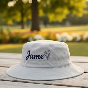 May include: A light gray bucket hat with the name "Jame" embroidered in navy blue script, along with a navy blue lobster graphic. The hat is sitting on a wooden surface.