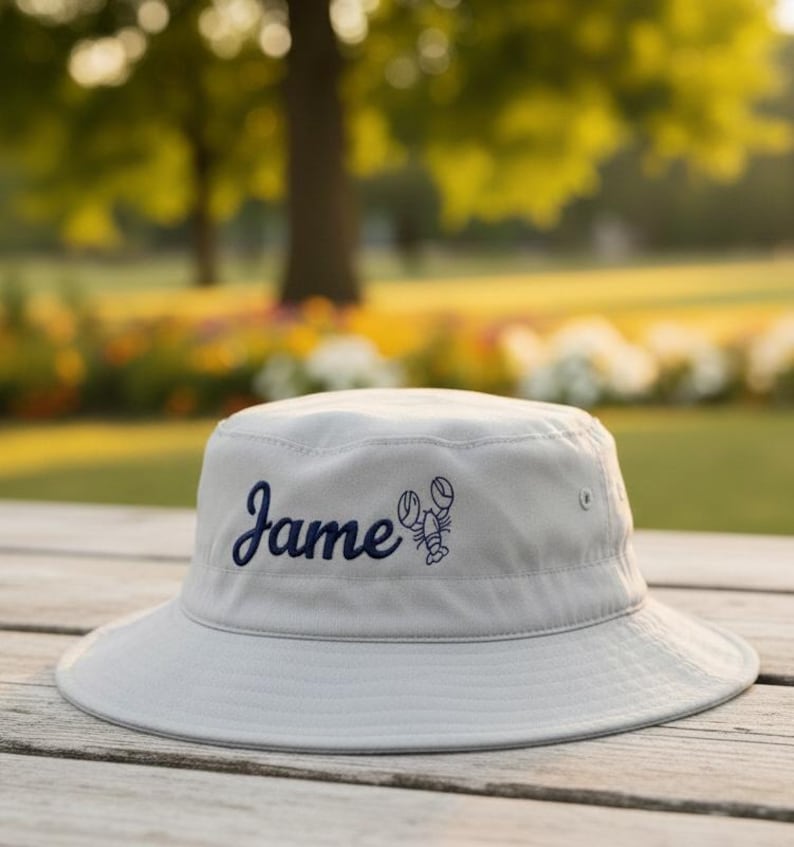 May include: A light gray bucket hat with the name "Jame" embroidered in navy blue script, along with a navy blue lobster graphic. The hat is sitting on a wooden surface.