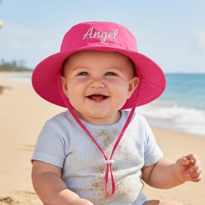 May include: A smiling baby wearing a bright pink sun hat with the word "Angel" embroidered on the front. The hat has a wide brim and a chin strap. The baby is wearing a light blue striped shirt and is sitting on a sandy beach.