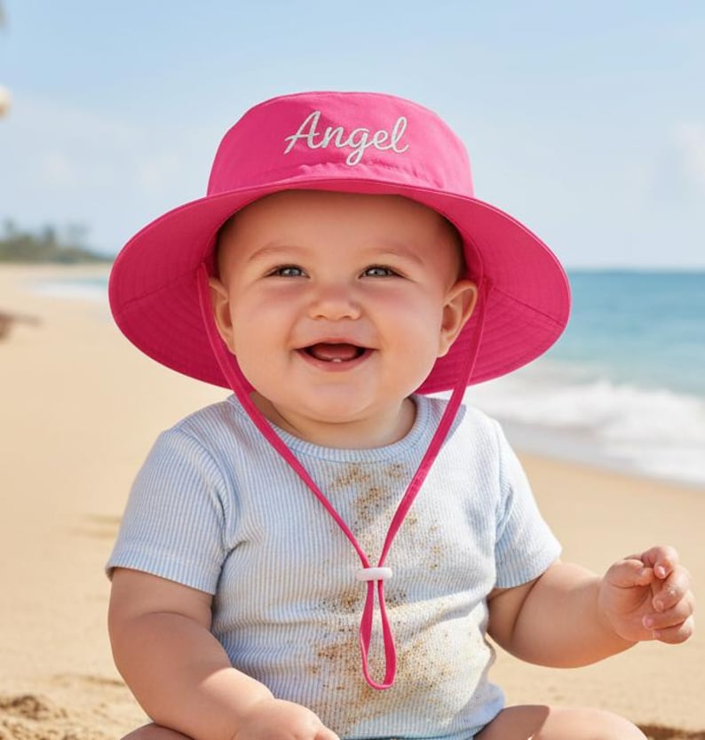 May include: A smiling baby wearing a bright pink sun hat with the word "Angel" embroidered on the front. The hat has a wide brim and a chin strap. The baby is wearing a light blue striped shirt and is sitting on a sandy beach.