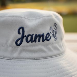 May include: Light gray bucket hat with the name "James" embroidered in navy blue script, accompanied by a navy blue lobster graphic. The hat is made of a textured fabric and is suitable for outdoor wear.