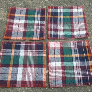 May include: Four square quilted coasters with a red, green, blue, and white plaid pattern. The coasters have a decorative quilted design with a leaf pattern.