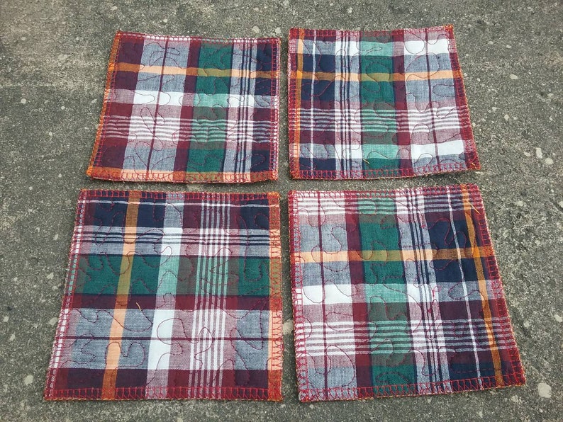 May include: Four square quilted coasters with a red, green, blue, and white plaid pattern. The coasters have a decorative quilted design with a leaf pattern.