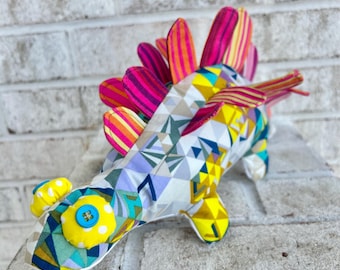 May include: A colorful, geometric patterned stuffed dinosaur toy with a bright yellow button eye and a pink, yellow, and orange striped back.